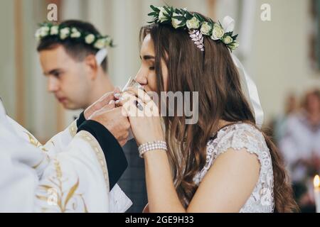 Elegante sposa e sposo in corone floreali bevendo Santa eucaristia durante il matrimonio in chiesa. Cerimonia nuziale in cattedrale. Matrimonio spirituale classico Foto Stock