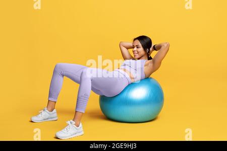 Donna nera sportiva che fa esercizi di abs sulla palla di fitness su sfondo giallo studio, spazio libero Foto Stock