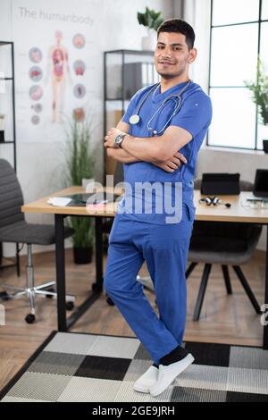 Bel giovane medico o studente di medicina sta guardando la macchina fotografica mentre si sta in piedi con le braccia ripiegate. Medico arabo maschile in posa con le braccia ripiegate in un moderno studio clinico leggero Foto Stock