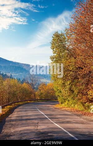 vecchia strada di campagna in montagna. alberi in fogliame colorato nella luce del mattino lungo la curva serpentina. tempo soleggiato con nuvole sul cielo sopra il d Foto Stock