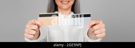 vista ritagliata di una donna sorridente offuscata che mostra carte di credito isolate su grigio, banner Foto Stock