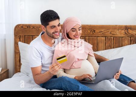 Felice coppia araba incinta shopping online a casa, utilizzando il computer portatile e la carta di credito, ottenendo pronto per la genitorialità Foto Stock