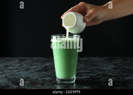 Mano femminile versando il latte in un bicchiere di latte matcha su sfondo nero Foto Stock
