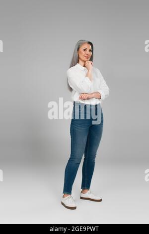 vista completa della donna matura in camicia bianca e dei jeans in piedi con la mano vicino al mento su sfondo grigio Foto Stock