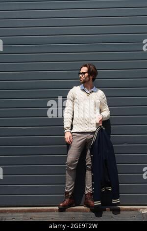 uomo alla moda in pullover bianco con giacca blu vicino alla parete grigia all'aperto Foto Stock