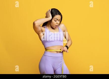 Shocked giovane donna nera che controlla il peso usando il metro a nastro sulla vita, tenendo la mano sulla testa, sfondo giallo Foto Stock