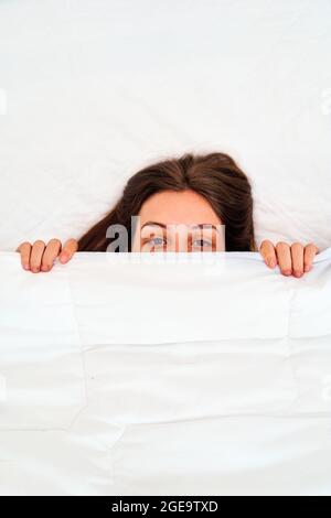 Dall'alto di giovane donna pigra guardando la macchina fotografica che copre metà del viso con morbida coperta bianca mentre si annidano in letto accogliente al mattino Foto Stock