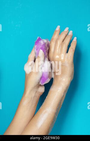 Mano di raccolto anonimo femmina spremitura spugna da bagno con schiuma bianca su sfondo blu Foto Stock