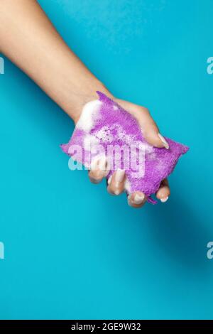 Mano di raccolto anonimo femmina spremitura spugna da bagno con schiuma bianca su sfondo blu Foto Stock