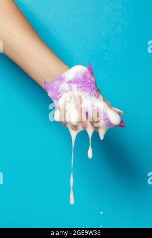 Mano di raccolto anonimo femmina spremitura spugna da bagno con schiuma bianca su sfondo blu Foto Stock