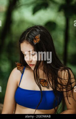 Tranquilla giovane femmina in bikini con farfalle ornamentali sul corpo e sui capelli in piedi contro il verde offuscato fogliame durante l'avventura estiva in Costa R. Foto Stock