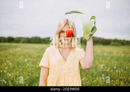 Delicato occhio di copertura femminile con fiore rosso tulipano mentre si sta in campo e guardando la fotocamera Foto Stock