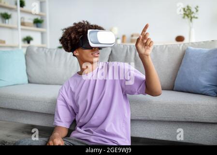 Ragazzo nero in visore VR che esplora la realtà virtuale a casa, tocca lo schermo immaginario, interagisce con il cyberspazio Foto Stock