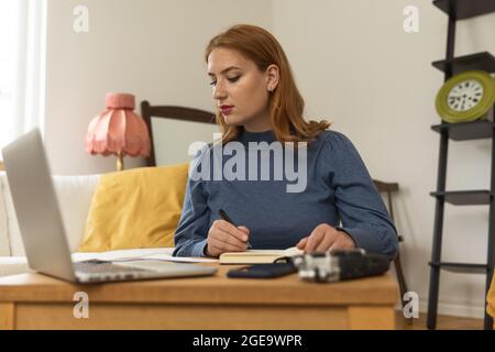 Host radio femminile focalizzato con microfono e cuffie che scrivono in blocco note durante la preparazione per la registrazione di podcast a casa Foto Stock