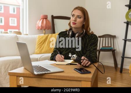 Host radio femminile attento e mirato con microfono e cuffie che scrivono in blocco note mentre si prepara per la registrazione di podcast a casa Foto Stock