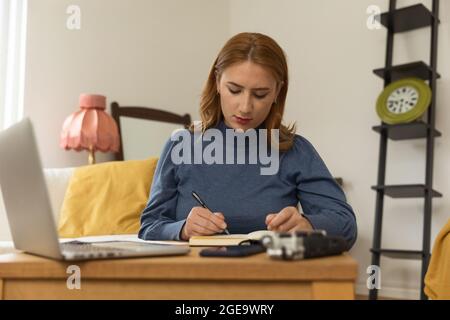 Host radio femminile focalizzato con microfono e cuffie che scrivono in blocco note durante la preparazione per la registrazione di podcast a casa Foto Stock