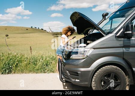 Giovane donna etnica con capelli afro che chiama per il servizio di riparazione su smartphone mentre si sta in piedi vicino camper van con cappuccio aperto sulla strada di campagna in estate Foto Stock