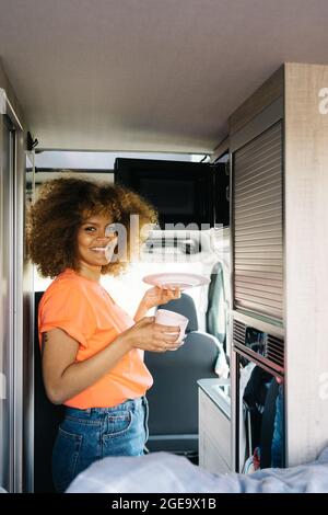 Vista laterale del viaggiatore femminile con piastra per capelli ricci nel forno a microonde mentre si riscaldano gli alimenti all'interno della moderna caravan Foto Stock