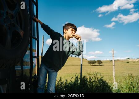 Carino piccolo viaggiatore mordendo gustoso sandwich e guardando via mentre appeso a scala di RV moderno in estate giorno in campagna Foto Stock