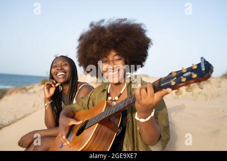 Felici le giovani amiche afro-americane che suonano la chitarra mentre si siedono insieme in riva al mare e si godono le vacanze estive Foto Stock