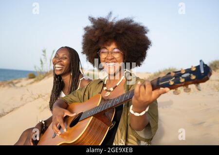 Felici le giovani amiche afroamericane che suonano la chitarra mentre si siedono con gli occhi chiusi insieme sulla spiaggia sabbiosa e si godono le vacanze estive Foto Stock