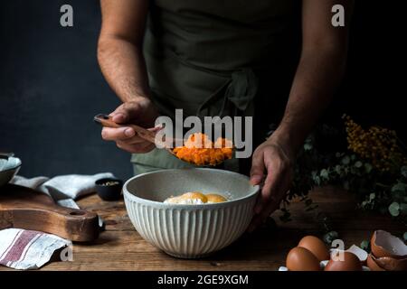 Cuoco maschile tagliato poco con cucchiaio di legno per mescolare la purea di zucca con le uova e la farina in ciotola mentre si prepara la torta sul tavolo di legno vicino al taglio Foto Stock