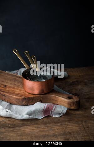 Pila di pentole di rame poste su tagliere di legno e asciugamani di lino su tavolo rustico su sfondo grigio Foto Stock