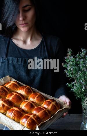 Panettiere femmina che tiene il puff appena sfornato a croce calda su vassoio da forno Foto Stock