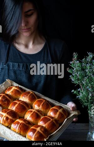 Panettiere femmina che tiene il puff appena sfornato a croce calda su vassoio da forno Foto Stock
