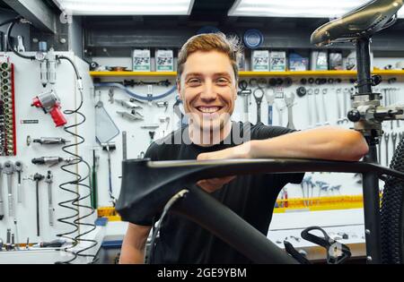 Felice meccanico maschio sorridente e guardando la macchina fotografica mentre si appoggia sulla bici sotto riparazione contro muro con attrezzi in garage Foto Stock