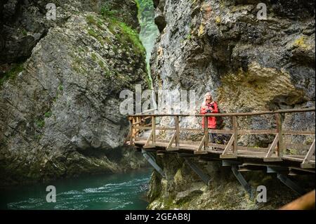 Giovane fotografo femminile in giacca colorata che percorre un sentiero lungo un fiume limpido attraverso la gola di Vintgar in Slovenia Foto Stock