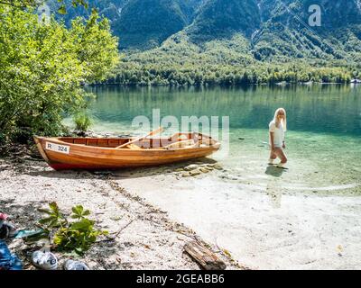 Donna bionda in piedi nel lago di Bohinj, accanto ad una barca sulla spiaggia Foto Stock