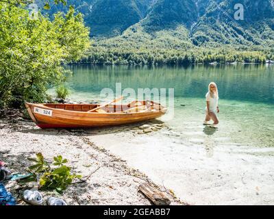 Donna bionda in piedi nel lago di Bohinj, accanto ad una barca sulla spiaggia Foto Stock