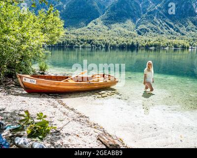 Donna bionda in piedi nel lago di Bohinj, accanto ad una barca sulla spiaggia Foto Stock