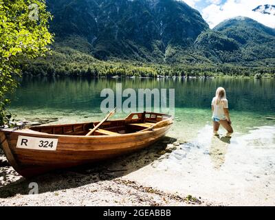 Donna bionda in piedi nel lago di Bohinj, accanto ad una barca sulla spiaggia Foto Stock