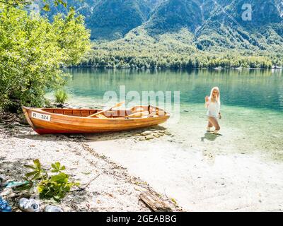 Donna bionda in piedi nel lago di Bohinj, accanto ad una barca sulla spiaggia Foto Stock