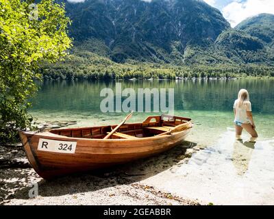 Donna bionda in piedi nel lago di Bohinj, accanto ad una barca sulla spiaggia Foto Stock