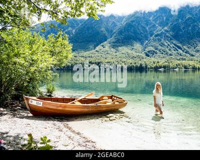 Donna bionda in piedi nel lago di Bohinj, accanto ad una barca sulla spiaggia Foto Stock