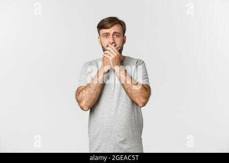 Immagine di un uomo spaventato in una t-shirt grigia, che brandisca e fremeva dalla paura, in piedi ansiosi su sfondo bianco Foto Stock