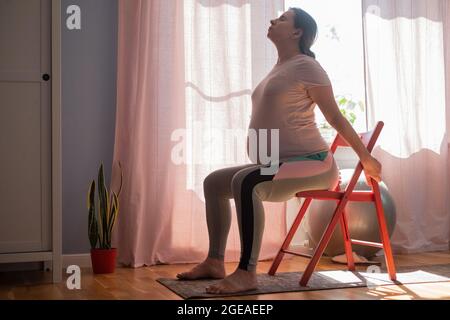 Donna incinta che si sta allenando all'interno praticando yoga a casa utilizzando la sedia Foto Stock