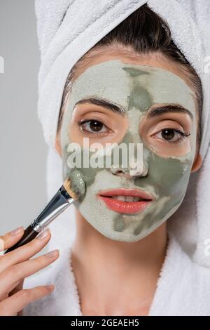 giovane donna con asciugamano sulla testa applicando maschera di argilla con spazzola cosmetica mentre guardando la fotocamera isolata su grigio Foto Stock