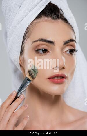 giovane donna con asciugamano sulla testa applicando maschera di argilla con spazzola cosmetica isolata su grigio Foto Stock