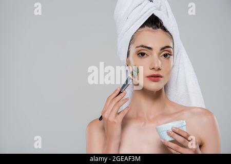 giovane donna con asciugamano sulla testa applicando maschera di argilla verde con spazzola cosmetica isolata su grigio Foto Stock