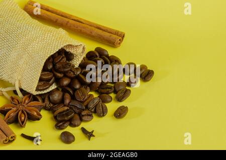 I chicchi di caffè sparsi in una piccola borsa si trovano con anice e cannella su uno sfondo giallo Foto Stock