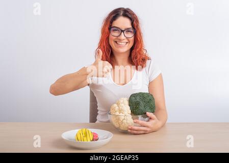 La donna caucasica preferisce cibo sano. Ragazza rossa sceglie tra broccoli e ciambelle su sfondo bianco. Foto Stock