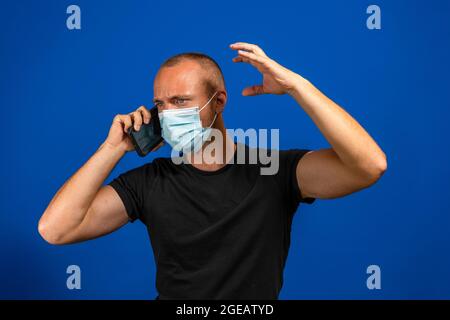 Giovane uomo caucasico parlando sul telefono cellulare molto arrabbiato, indossa una maschera chirurgica per proteggersi dal coronavirus. Isolato su studio blu Foto Stock