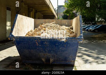 Dumpster blu con mattoni rotti e intonaco dopo riparazione appartamento in una giornata estiva soleggiata. Grande contenitore con detriti edili sulla strada. Foto Stock