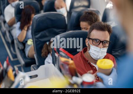 Assistente di volo che serve bevande al passeggero maschio in maschera protettiva a bordo. Viaggiare in aereo durante la pandemia di Covid19 Foto Stock