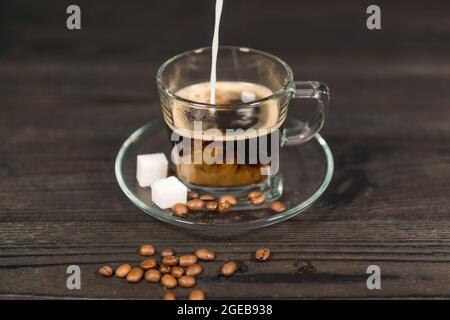 Una tazza di vetro, su un piattino, riempita con caffè estratto. Un flusso di latte bianco si versa nel caffè dall'alto. Foto Stock