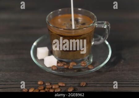 Una tazza di vetro, su un piattino, riempita con caffè estratto. Un flusso di latte bianco si versa nel caffè dall'alto. Foto Stock